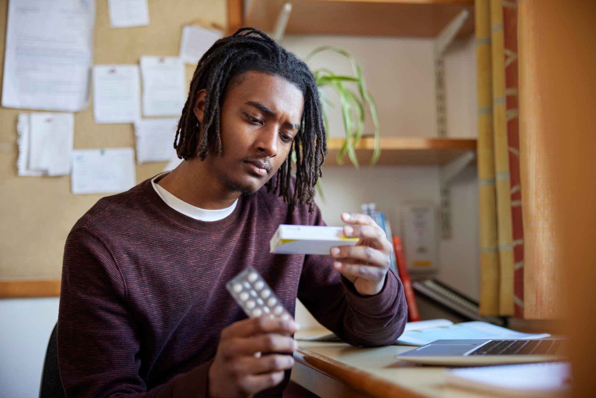 A young man reading the label on a box of medication while sat at a desk.