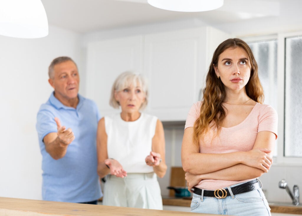 Parents lecturing their adult daughter at home while she crosses her arms and faces away from them.